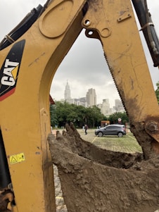 A large excavator arm with a muddy scoop is in the foreground, set against an urban backdrop. Skyscrapers and a cloudy sky can be seen in the background. A parked car and a few pedestrians are visible near some greenery, giving a contrast between construction and city life.