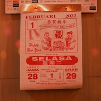 A calendar page from February 1, 2022, featuring red and white colors. It includes greetings for the Chinese New Year, with text in multiple languages including Bahasa Indonesia. The design features two cartoon figures holding items and traditional Chinese motifs, such as fireworks and intricate patterns.