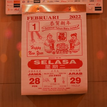 A calendar page from February 1, 2022, featuring red and white colors. It includes greetings for the Chinese New Year, with text in multiple languages including Bahasa Indonesia. The design features two cartoon figures holding items and traditional Chinese motifs, such as fireworks and intricate patterns.