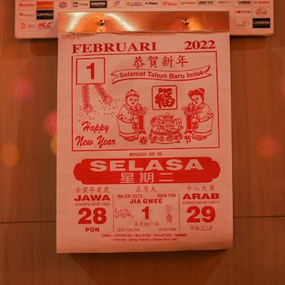 A calendar page from February 1, 2022, featuring red and white colors. It includes greetings for the Chinese New Year, with text in multiple languages including Bahasa Indonesia. The design features two cartoon figures holding items and traditional Chinese motifs, such as fireworks and intricate patterns.