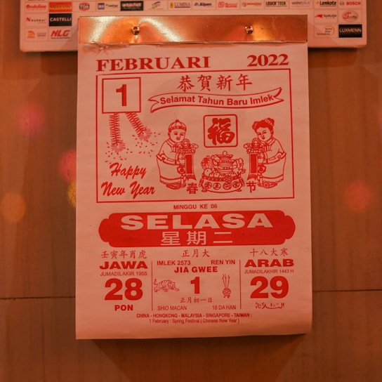 A calendar page from February 1, 2022, featuring red and white colors. It includes greetings for the Chinese New Year, with text in multiple languages including Bahasa Indonesia. The design features two cartoon figures holding items and traditional Chinese motifs, such as fireworks and intricate patterns.