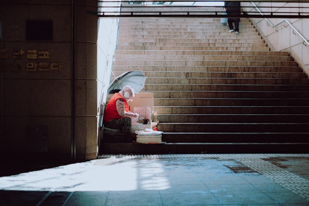 An elderly person in a red vest is sitting at the bottom of a staircase. They have an umbrella and a variety of small items, possibly for sale, around them. The setting appears to be an underpass or subway entrance with muted lighting and concrete surroundings.