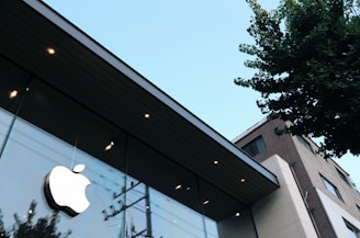 A modern building with sleek glass windows and a prominent Apple logo. The structure is angled upwards, suggesting a tall facade. In the background, a clear blue sky is visible, and nearby, a tree casts shadows on the glass. The building design appears contemporary, with smooth lines and minimalistic architecture.