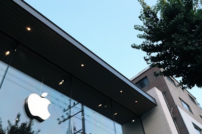A modern building with sleek glass windows and a prominent Apple logo. The structure is angled upwards, suggesting a tall facade. In the background, a clear blue sky is visible, and nearby, a tree casts shadows on the glass. The building design appears contemporary, with smooth lines and minimalistic architecture.
