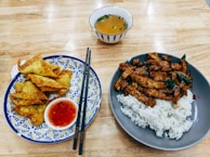 A wooden table features two plates of food. The left plate has crispy fried wontons accompanied by a small dish of red chili sauce, with black chopsticks placed across the plate. The right plate contains a serving of white rice topped with stir-fried pieces of meat and vegetables. In the background, a small bowl with soup garnished with chopped greens is visible.