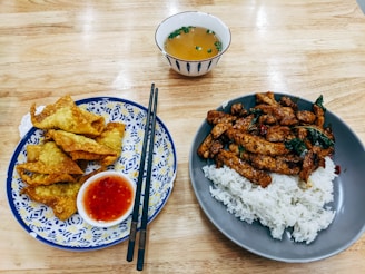 A wooden table features two plates of food. The left plate has crispy fried wontons accompanied by a small dish of red chili sauce, with black chopsticks placed across the plate. The right plate contains a serving of white rice topped with stir-fried pieces of meat and vegetables. In the background, a small bowl with soup garnished with chopped greens is visible.
