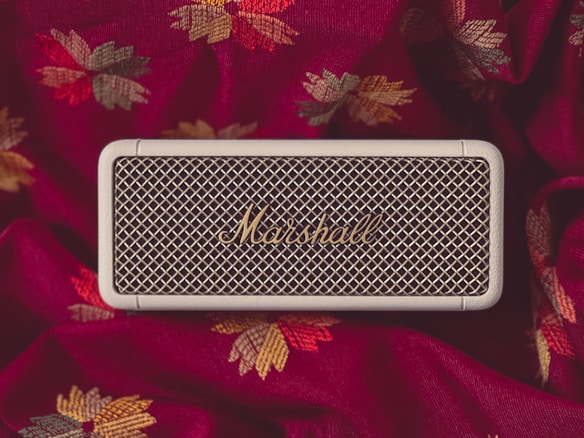 A portable speaker with a vintage design is placed on a richly colored fabric with an intricate leaf pattern. The speaker has a mesh front and bears the brand name 'Marshall' in gold letters.