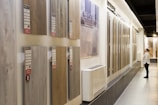 A showroom displaying various wooden flooring samples mounted on walls. A person is observing the samples on the right side, wearing casual attire. The samples are labeled with prices and descriptions. The room is well-lit with a modern design and uses neutral tones.
