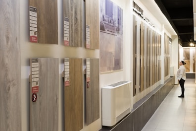 A showroom displaying various wooden flooring samples mounted on walls. A person is observing the samples on the right side, wearing casual attire. The samples are labeled with prices and descriptions. The room is well-lit with a modern design and uses neutral tones.