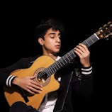 A person is playing an acoustic guitar with focus and concentration. The guitar has a wooden finish with intricate detailing around the sound hole. The person is wearing a black jacket with white stripes on the sleeves and appears to be in a dimly lit environment, highlighted against a dark background.