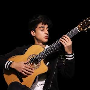 A person is playing an acoustic guitar with focus and concentration. The guitar has a wooden finish with intricate detailing around the sound hole. The person is wearing a black jacket with white stripes on the sleeves and appears to be in a dimly lit environment, highlighted against a dark background.