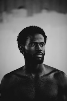A black and white photograph of a shirtless man with short curly hair and facial hair, gazing off to the side against a blurred background.