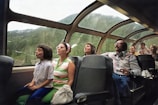 A group of people are seated in a train coach with large panoramic windows. They are gazing out at a scenic view, likely of mountains and forests. The seats are arranged in rows, and the passengers include adults and children, all appearing relaxed and engaged with the scenery.