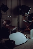 A dimly lit room filled with photography equipment, including a brown leather chair, a director's chair, a softbox light, and a silver reflector placed on the carpeted floor.