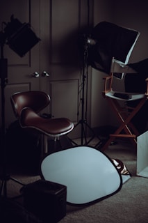 A dimly lit room filled with photography equipment, including a brown leather chair, a director's chair, a softbox light, and a silver reflector placed on the carpeted floor.