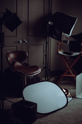 A dimly lit room filled with photography equipment, including a brown leather chair, a director's chair, a softbox light, and a silver reflector placed on the carpeted floor.