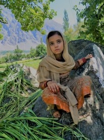 A young child wearing traditional clothing is sitting on a large rock surrounded by greenery and trees in a mountainous landscape. The child appears to be looking at the camera with a calm expression. The background features lush fields and tall mountains under a clear blue sky.