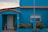 A vibrant blue house with terra cotta roof tiles features a small, gated front porch with a white door. A metal fence surrounds the porch area, adorned with lush green plants with yellow flowers. A pink flamingo statue stands near the entrance, adding a playful touch.