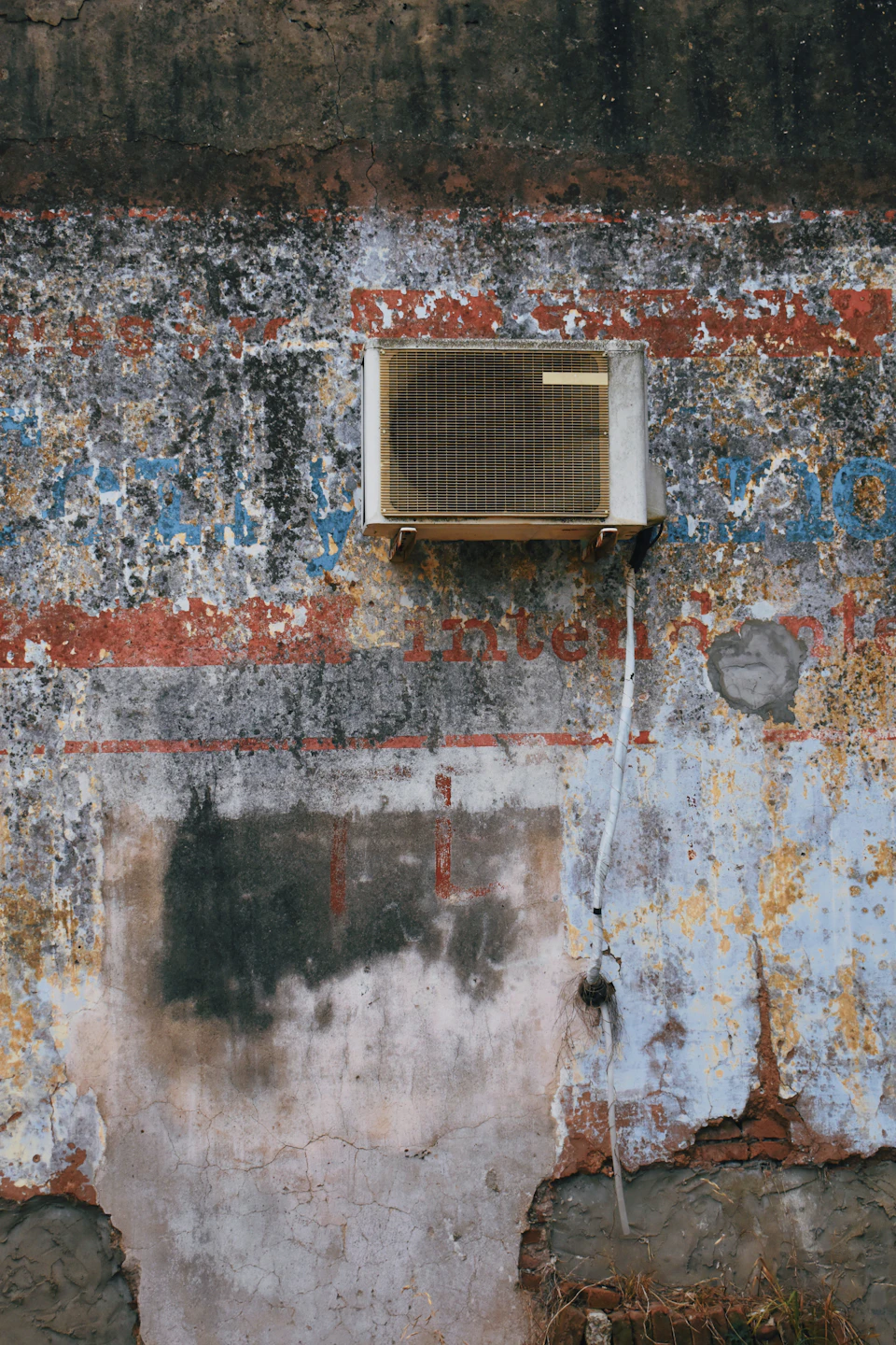 The image shows a weathered concrete wall with peeling paint and patches of rust, giving it an aged appearance. A white air conditioning unit is mounted on the right side of the wall, facing towards the left side of the frame. The wall has a rough texture, adding to its worn-out look. In the bottom right corner, there's a small piece of paper with writing that appears to be graffiti or some other form of vandalism.
