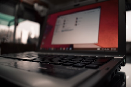 A laptop with an open screen displaying a blurred interface. The focus is on the keyboard, with keys in sharp detail. The background appears blurry, suggesting an indoor setting with some natural light coming in through a window.