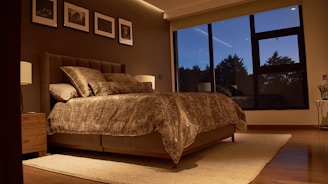 Спалня с голямо легло и голям прозорец през нощта, нежно осветена от лампи върху шкафове, с кафява 