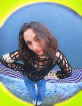 A woman with shoulder-length hair and a confident expression is wearing a black, open-knit sweater with cream detailing over a blue top. She poses with her hands on her hips. Her jeans are light blue, and she wears white shoes. The background features a vivid circular frame in shades of green, blue, and yellow, and a purple striped pattern on the floor.