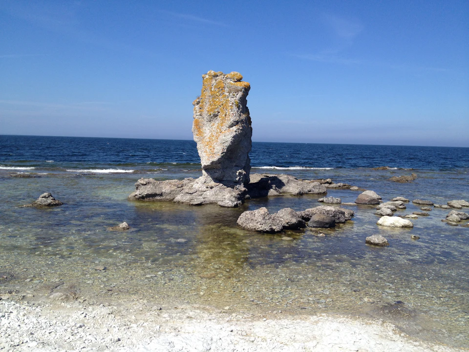 The image depicts a rocky outcropping with a large, moss-covered rock formation situated on a beach. The rock formation is surrounded by small rocks and pebbles, creating a rugged landscape. The clear blue sky above contrasts with the greenish hue of the water below, which appears to be calm and serene.