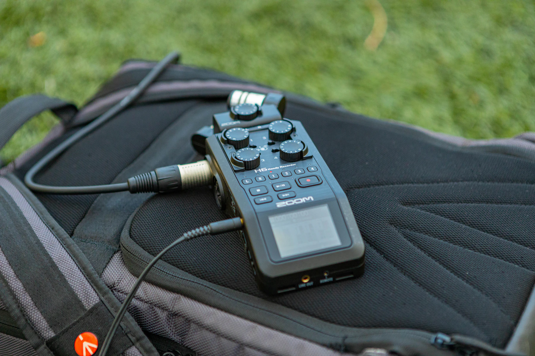 A handheld audio recorder with a cable plugged in, resting on a backpack on grass