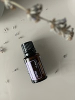 A small dark brown glass bottle labeled 'Patchouli' is pictured on a smooth, neutral background. A stem with dried lavender flowers is blurred in the foreground, with a few scattered lavender buds around the bottle.