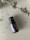 A small dark brown glass bottle labeled 'Patchouli' is pictured on a smooth, neutral background. A stem with dried lavender flowers is blurred in the foreground, with a few scattered lavender buds around the bottle.