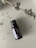 A small dark brown glass bottle labeled 'Patchouli' is pictured on a smooth, neutral background. A stem with dried lavender flowers is blurred in the foreground, with a few scattered lavender buds around the bottle.