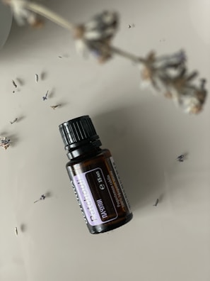 A small dark brown glass bottle labeled 'Patchouli' is pictured on a smooth, neutral background. A stem with dried lavender flowers is blurred in the foreground, with a few scattered lavender buds around the bottle.
