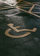 A painted yellow symbol on a textured asphalt surface representing a wheelchair-accessible parking space. The background shows part of a grid of yellow lines indicating the parking area.