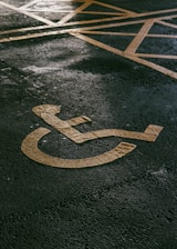A painted yellow symbol on a textured asphalt surface representing a wheelchair-accessible parking space. The background shows part of a grid of yellow lines indicating the parking area.