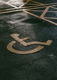 A painted yellow symbol on a textured asphalt surface representing a wheelchair-accessible parking space. The background shows part of a grid of yellow lines indicating the parking area.