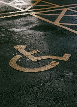 A painted yellow symbol on a textured asphalt surface representing a wheelchair-accessible parking space. The background shows part of a grid of yellow lines indicating the parking area.