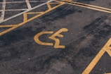 A parking lot features a yellow painted symbol on the ground indicating a reserved space for disability access. The asphalt is dark and slightly worn, with parallel yellow lines creating a distinct parking area.