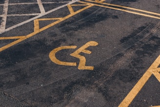 A parking lot features a yellow painted symbol on the ground indicating a reserved space for disability access. The asphalt is dark and slightly worn, with parallel yellow lines creating a distinct parking area.