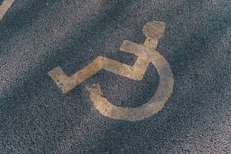 A faded yellow wheelchair symbol is painted on a textured asphalt surface, often seen in parking areas reserved for individuals with disabilities.