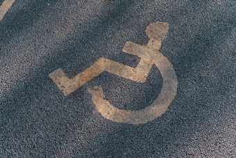 A faded yellow wheelchair symbol is painted on a textured asphalt surface, often seen in parking areas reserved for individuals with disabilities.