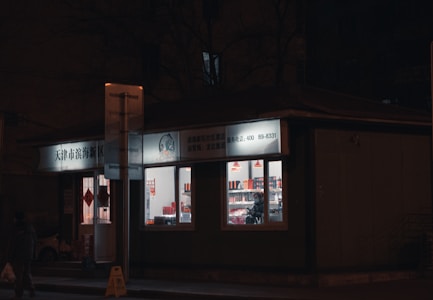 A small shop is brightly lit against a dark street. The windows reveal shelves inside filled with various products, and a person standing inside. The signage above the store is in Chinese characters, and there are red hanging lamps visible inside.