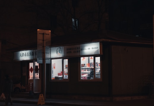 A small shop is brightly lit against a dark street. The windows reveal shelves inside filled with various products, and a person standing inside. The signage above the store is in Chinese characters, and there are red hanging lamps visible inside.
