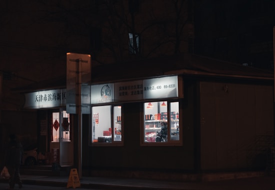 A small shop is brightly lit against a dark street. The windows reveal shelves inside filled with various products, and a person standing inside. The signage above the store is in Chinese characters, and there are red hanging lamps visible inside.