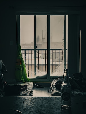 A dimly lit room with a large sliding glass door leading to a balcony, where droplets of water are visible on the glass. Inside, a bright yellow-green jacket hangs to the left of the door. The floor is covered with mattresses or blankets, and a laptop is open on one of them. A glass bottle with a label stands on a table in the foreground.