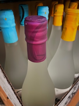 Bottles topped with colorful wax seals are neatly arranged, featuring hues of blue, purple, and yellow. The bottles appear frosted or opaque, possibly indicating alcoholic or artisanal beverages. The tops and arrangement suggest a display on a shelf possibly in a store or winery.