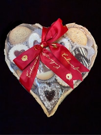 A heart-shaped basket filled with an assortment of cookies, adorned with a red ribbon tied into a bow. The cookies vary in style, featuring jam-filled centers, powdered sugar coatings, and chocolate drizzles.