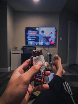 A person is lounging in a living room with legs stretched out while holding a partially unwrapped chocolate bar. A television is on in the background displaying a paused streaming service menu and a blurred image of a show or movie.