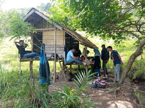 A group of people are gathered around a small, rustic wooden hut in a lush, green outdoor setting. Some individuals appear to be relaxing, while others are engaged in conversation or activities under the shade of trees.