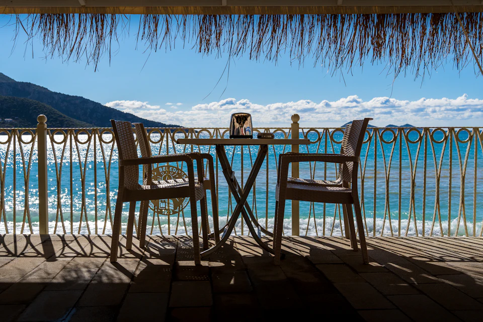 The image depicts a serene beachfront patio overlooking the ocean. The patio is furnished with two brown wicker chairs, each accompanied by a small table and a white umbrella. The chairs are positioned facing the ocean, providing an unobstructed view of the vast expanse of water. A metal railing runs along the edge of the patio, adding to its rustic charm. In the background, there's a hill covered in lush green trees that adds depth to the scene.

The sky above is a clear blue with scattered clouds, suggesting a bright and sunny day. The ocean stretches out towards the horizon, its surface shimmering under the sunlight.