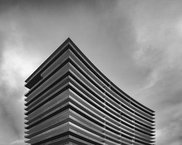 A modern, multi-story building with a sleek, curved design set against a partly cloudy sky. The architecture features horizontal lines and large glass windows across each floor, emphasizing a contemporary style.